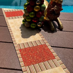 May include: A beige and red Christmas table runner with a striped border and two red rectangular sections with gold stars. A decorative Christmas tree and a gold reindeer are in the background.