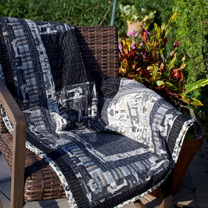 May include: A black and white patterned quilt with a sewing theme is draped over a brown wicker chair. The chair is positioned near a swimming pool and green foliage.