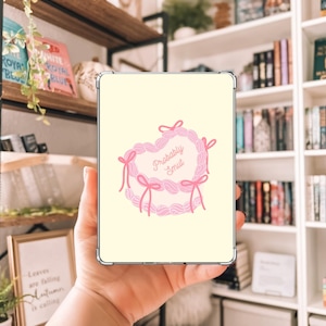 May include: A light yellow tablet case featuring a pink heart-shaped design with the words "Probably Smut" in a cursive font. The heart is decorated with pink bows and a scalloped border.