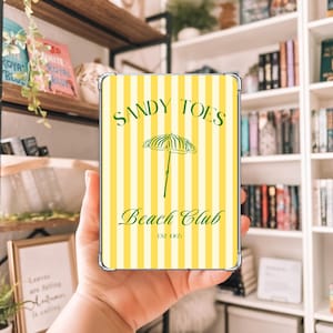Puede incluir: Una funda transparente para teléfono con un diseño de rayas amarillas y blancas. La funda presenta el texto "Sandy Toes Beach Club" en verde, junto con un gráfico de sombrilla verde. También incluye el texto "EST 1905".