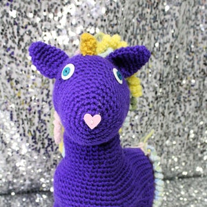 May include: A purple crocheted unicorn toy with a yellow and white mane and tail. The unicorn has blue eyes and a pink heart nose.