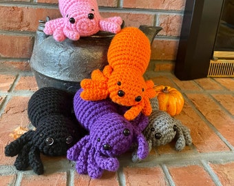 May include: A collection of five crocheted spider plushies in pink, orange, purple, black, and gray, each featuring eight legs and two black plastic eyes. The spiders are arranged around a black metal kettle and a small orange decorative pumpkin.