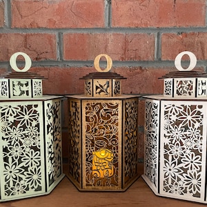 Könnte beinhalten: Drei Holzlaternen mit filigranen Blumendesigns. Die Laternen sind sechseckig und haben eine hellbraune Oberfläche. Die Laternen sind mit einem warmweißen Licht beleuchtet. Die Laternen sind mit dem Buchstaben "O" beschriftet.