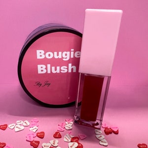 Könnte beinhalten: Ein rosa und schwarzes Glas mit dem Text "Bougie Blush By Jay" und ein rosa und klares Röhrchen mit roter flüssiger Rouge.