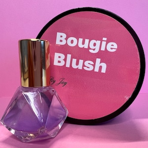 Peut inclure: Un flacon en verre transparent avec un liquide violet et un bouchon doré. Le flacon a la forme d'un diamant. Le flacon est posé sur une surface rose. L'étiquette sur le flacon indique "Bougie Blush by Jay".