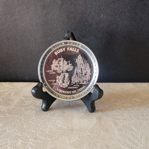 May include: A silver-colored round metal coaster with a black stand. The coaster features a black and white image of Ruby Falls, Lookout Mt. Tenn. The text "Ruby Falls" is printed in black on the coaster.