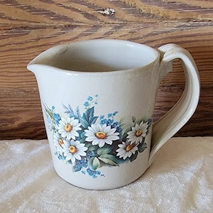 May include: A white ceramic pitcher with a floral design featuring white daisies and blue forget-me-nots. The pitcher has a wide, rounded base and a handle.