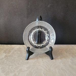 Peut inclure: Une assiette en verre transparent avec un bord perlé, présentée sur un support noir. L'assiette a une surface texturée et un design circulaire.