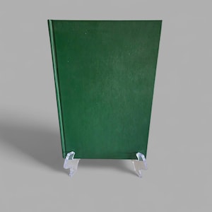 May include: A hardcover book with a dark green cover, displayed on a clear plastic stand. The book's spine is visible, and the cover has a slightly textured appearance. The background is a solid, neutral gray.
