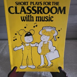 May include: A vintage yellow book titled "Short Plays for the Classroom with music." The cover features cartoon illustrations of two children in mouse costumes, with musical notes. The book is propped up on a wooden stand.