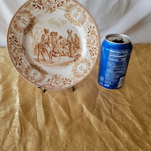 May include: A brown and white ceramic plate with a decorative border and a scene of soldiers in a battle. The plate has the text "Bataille de Montreal" and "2 Juillet 1760" on it.