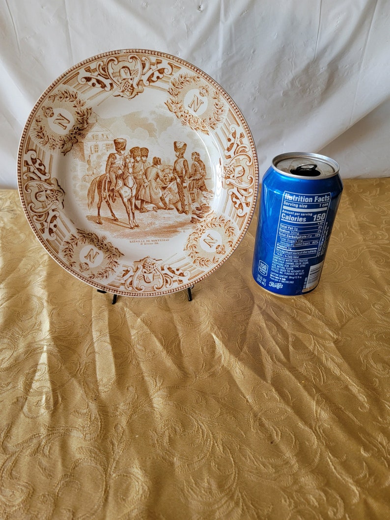 May include: A brown and white ceramic plate with a decorative border and a scene of soldiers in a battle. The plate has the text "Bataille de Montreal" and "2 Juillet 1760" on it.
