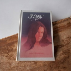 Könnte beinhalten: Ein weißes Taschenbuch mit dem Titel "Hagar, A Novel" in kursivschrift. Der Umschlag zeigt ein Gemälde einer Frau mit langen dunklen Haaren vor einem rosafarbenen Hintergrund.