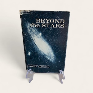 Peut inclure: Un livre vintage intitulé "Beyond the Stars" avec une couverture bleu foncé représentant une image de galaxie. Le livre est maintenu par un support en plastique transparent. Le nom de l'auteur, Bobby Jackson, est imprimé en bas.