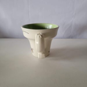 May include: Cream-colored ceramic mug with a green interior and a handle. The mug has a flared shape and features bee illustrations and the text "the honey bee". A decorative and functional item for beverages.