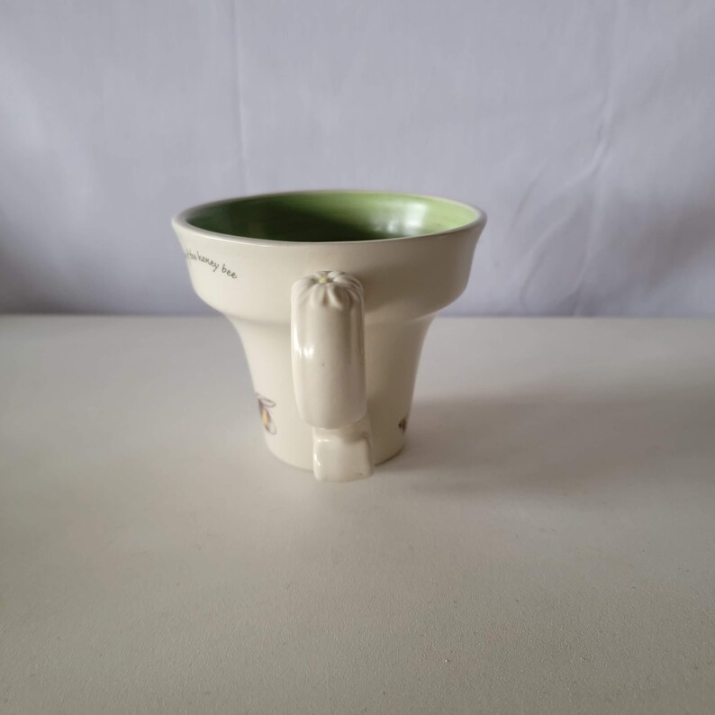 May include: Cream-colored ceramic mug with a green interior and a handle. The mug has a flared shape and features bee illustrations and the text "the honey bee". A decorative and functional item for beverages.
