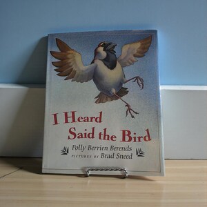 Może przedstawiać: Książka dla dzieci zatytułowana "I Heard Said the Bird" z kolorową ilustracją wróbla w locie. Okładka książki zawiera nazwiska autora i ilustratora i jest wyświetlana na metalowym stojaku na książki.