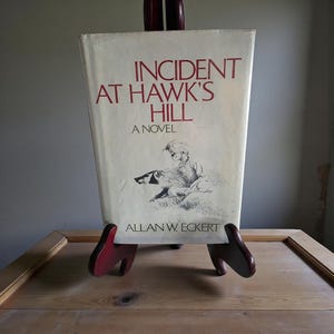 May include: A vintage book titled "Incident at Hawk's Hill: A Novel" by Allan W. Eckert, displayed on a wooden easel. The book cover features a drawing of a person with a badger. The title and author's name are in red.