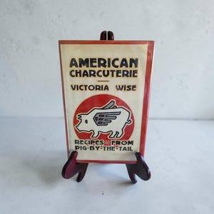 May include: A rectangular cookbook titled American Charcuterie by Victoria Wise, featuring a graphic of a winged pig inside a red circle. The book cover is cream-colored with black text and red accents, displayed upright on a dark brown wooden stand.