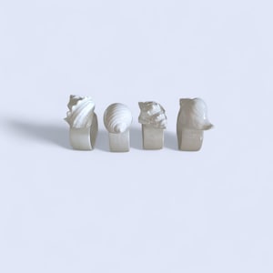 May include: Four off-white ceramic napkin rings, each with a unique seashell design. The rings are in the shape of a band with a seashell sculpture on top. The seashells include a spiral, a conch, and other textured shapes.