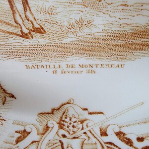 May include: A white ceramic plate with a brown image of a battle scene. The text "Bataille de Montereau 18 fevrier 1814" is printed on the plate.
