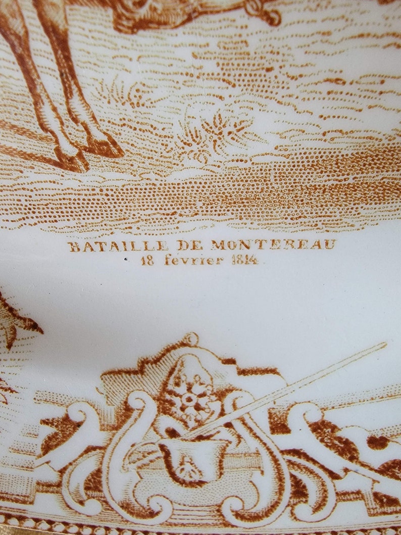 May include: A white ceramic plate with a brown image of a battle scene. The text "Bataille de Montereau 18 fevrier 1814" is printed on the plate.
