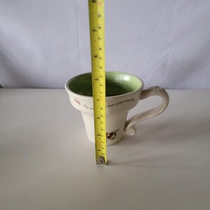 May include: Cream-colored ceramic mug with a green interior and a decorative bee design. The mug is approximately 4 inches tall. The handle is a unique, curved shape. Text on the mug reads "The world is the hum of the honey bee."