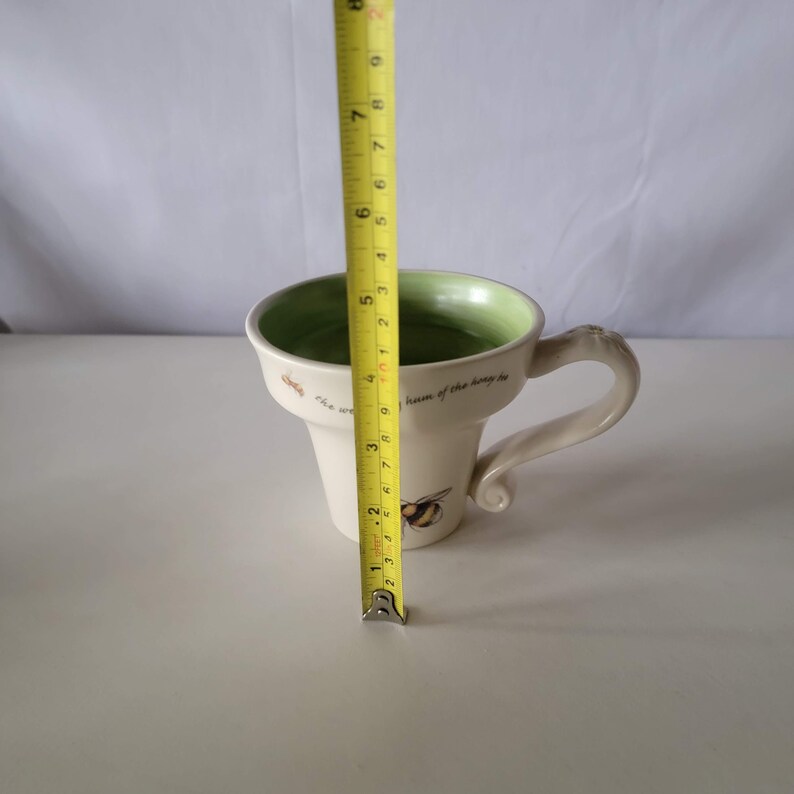May include: Cream-colored ceramic mug with a green interior and a decorative bee design. The mug is approximately 4 inches tall. The handle is a unique, curved shape. Text on the mug reads "The world is the hum of the honey bee."