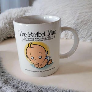 May include: White ceramic coffee mug with a cartoon baby on it. The mug says "The Perfect Man (He's young. He's cute. And he'll always come crawling back to you.)"