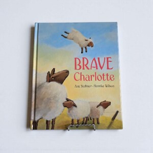 May include: A children's book titled "Brave Charlotte" with illustrations of sheep. The cover features a sheep jumping in the air against a blue sky, with other sheep grazing below. The book is by Anu Stohner and Henrike Wilson.