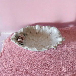 以下が含まれることがあります： ピンクの花のアクセントが付いた、葉の形をした白いセラミック製の皿。皿はピンクのふわふわした表面の上に置かれています。