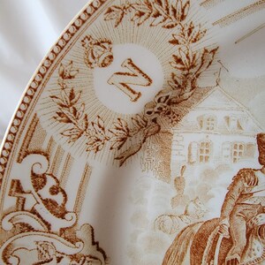 May include: A white ceramic plate with a brown design. The plate features a crown, the letter "N", a building, and a person on horseback. The design is reminiscent of a historical scene.