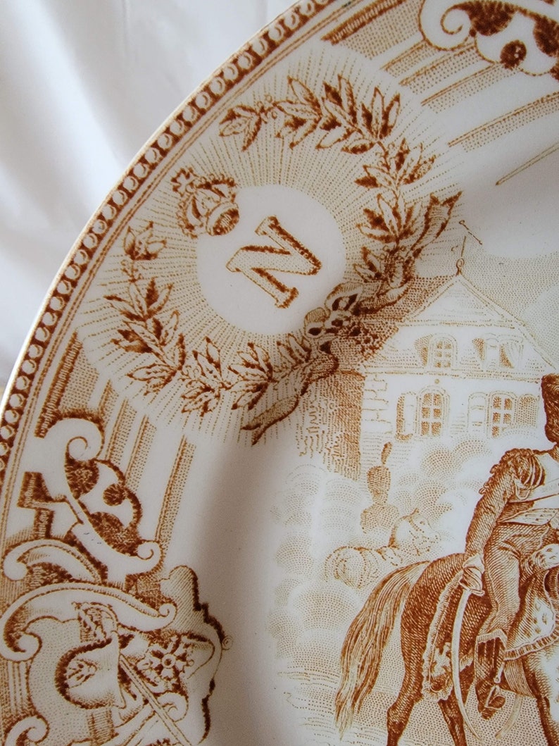 May include: A white ceramic plate with a brown design. The plate features a crown, the letter "N", a building, and a person on horseback. The design is reminiscent of a historical scene.