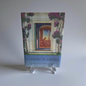 May include: A book titled "A House Blessing" by Welleran Poltarnees. The cover art features a painting of a doorway with a person inside, framed by pink flowering trees. The book is displayed on a clear stand.