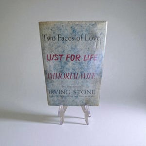 May include: A vintage book titled "Two Faces of Love" with the sub-titles "Lust for Life" and "Immortal Wife" by Irving Stone. The book has a blue and white cover and is displayed on a clear plastic stand.
