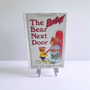 May include: A children's book titled "The Bear Next Door" with illustrations of two bears. The cover features the text "The Excellent!" in red, with the title in a larger font. The book is displayed on a clear plastic stand.
