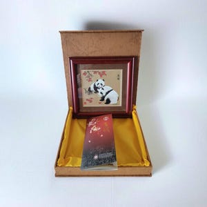 May include: A framed artwork featuring two pandas in a natural setting, presented in a decorative box. The artwork is in a dark wood frame, and the box has a gold satin lining. A brochure with red and gold accents is also included.