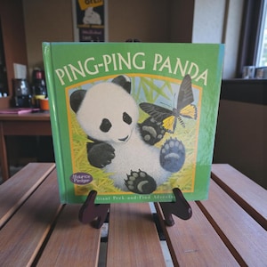 May include: A children's book titled "Ping-Ping Panda" is displayed on a wooden table. The cover features a panda bear illustration with a butterfly. The book is green with the title in large, white letters. The book is held by a dark purple book stand.