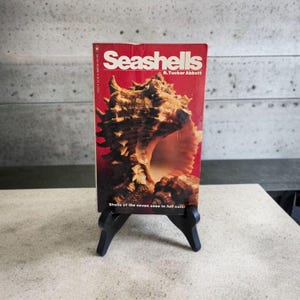 Peut inclure: Un livre vintage intitulé "Seashells" de R. Tucker Abbott. La couverture est rouge avec une grande illustration de coquillage. Le livre est présenté sur un petit chevalet noir. Le texte "Shells of the seven seas in full color" est également visible.