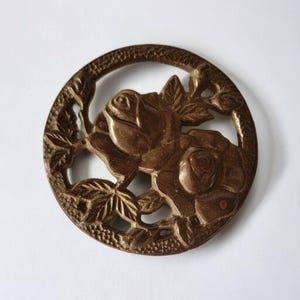 Puede incluir: Un adorno circular de latón con un diseño detallado de dos rosas y hojas. Las rosas están en relieve y las hojas están recortadas, creando un patrón delicado e intrincado.