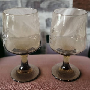 May include: Two clear glass wine glasses with a brown tint and etched floral designs. The glasses have a round bowl and a stem.