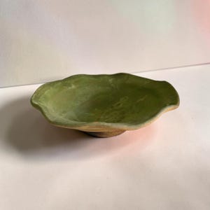 Puede incluir: Un cuenco de cerámica circular y poco profundo con un borde ondulado y festoneado. El interior presenta un esmalte verde salvia moteado con sutiles variaciones de tono, mientras que la base exterior revela una textura de arcilla marrón natural.