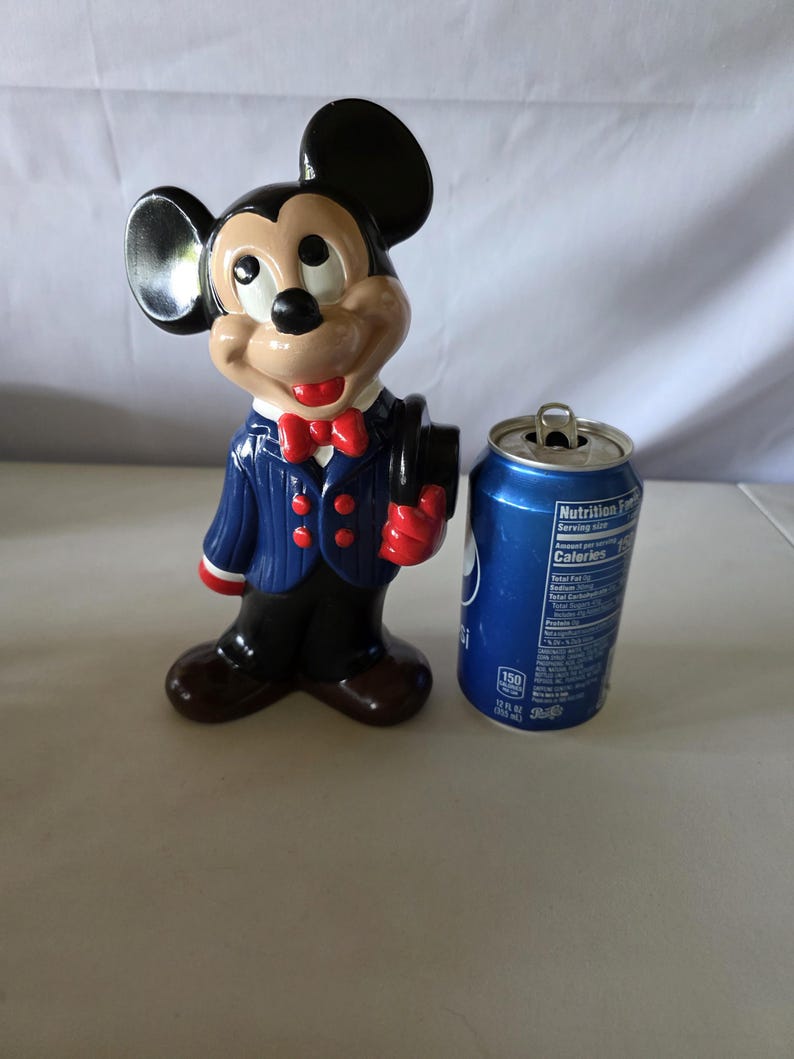 K&ouml;nnte beinhalten: Eine Keramikfigur von Mickey Mouse in einem blau gestreiften Anzug, roter Fliege und schwarzem Zylinder. Daneben steht eine blaue Pepsi-Dose.