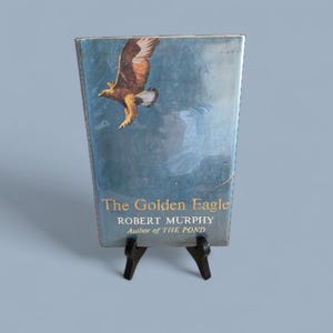 Puede incluir: Un libro antiguo titulado "The Golden Eagle" de Robert Murphy, expuesto sobre un pequeño soporte negro. La portada presenta una pintura de un águila real en vuelo sobre un cielo azul. El libro está ligeramente desgastado.