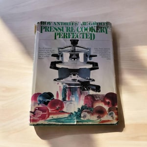 Pode incluir: Um livro de cozinha branco com um título verde, "Roy Andries De Groot Pressure Cookery Perfected". A capa apresenta uma panela de pressão prateada com vegetais e um pimentão vermelho.