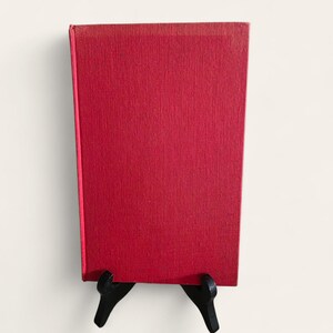 May include: A red hardcover book with a textured cover, displayed on a black easel. The book's spine is visible, and the cover is a solid, vibrant red. The book is set against a white background.