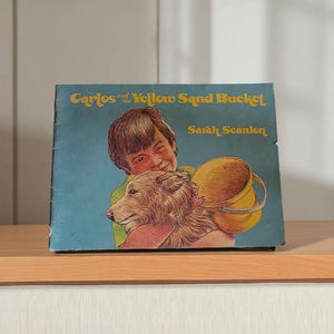 May include: A vintage children's book titled "Carlos and the Yellow Sand Bucket" by Sarah Scanlon. The cover features an illustration of a child hugging a dog, holding a yellow sand bucket. The book is set against a blue background.