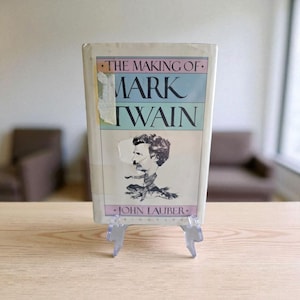 Może przedstawiać: Książka w stylu vintage zatytułowana "The Making of Mark Twain" z portretem Marka Twaina na okładce. Książka jest prezentowana na przezroczystym plastikowym stojaku, z jasnym tłem.