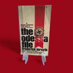 May include: A vintage paperback book titled "The Odessa File" by Frederick Forsyth. The cover features a red and beige design with the title in bold black letters and a Nazi emblem. The book is displayed on a clear plastic stand.