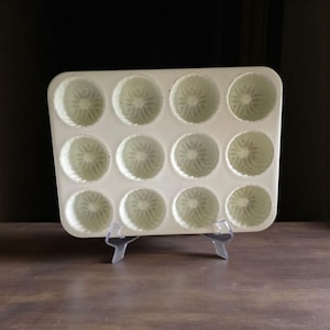 May include: A white, rectangular baking mold with 12 circular indentations. Each indentation has a detailed, floral-like pattern. The mold is displayed on a clear plastic stand against a dark background, suggesting a kitchen or baking theme.